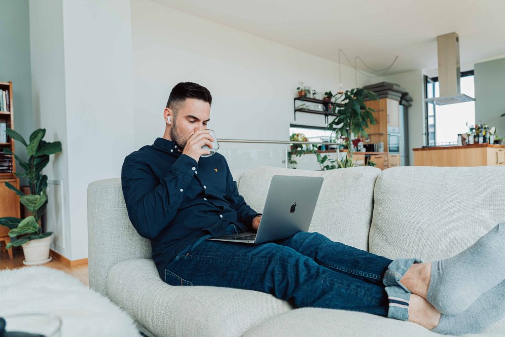 A man is seated comfortably on his couch with a laptop across his legs and sipping from a mug