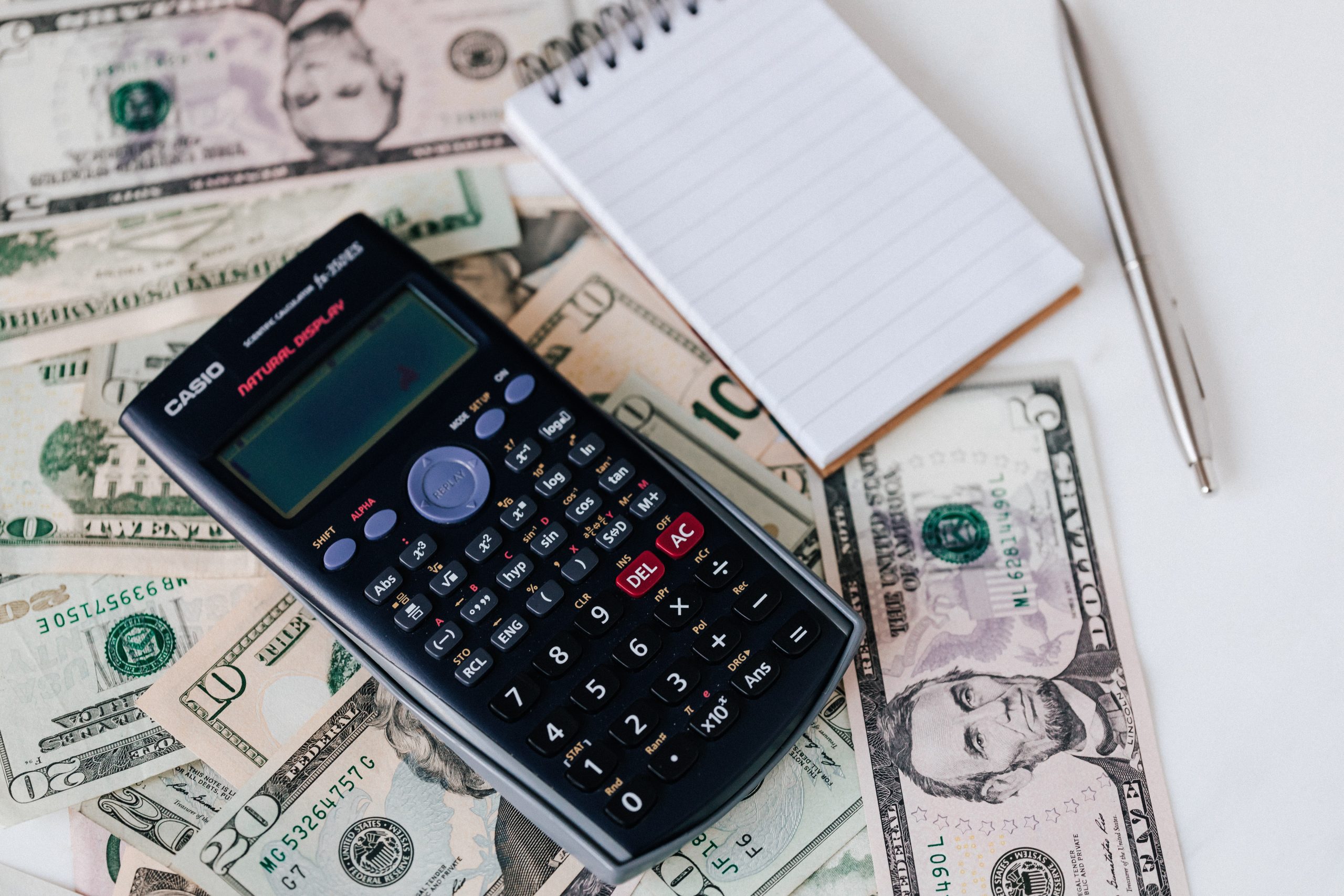 A calculator rests on top of several stacks of currency next to a notebook